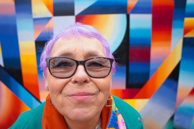 A woman with purple hair and glasses standing in front of a colorful wall